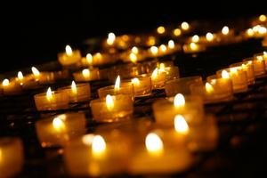 Brennende Kerzen vor dunklem Hintergrund. Etliche brennende Kerzen sind im Bild zu sehen. Die Kerzen im Vorder- und Hintergrund verlaufen in der Unschärfe.