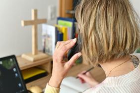 Pfarrerin Nele Schomakers in einem Büro beim Telefonieren - seitlich von hinten aufgenommen mit Blick auf den Bürotisch.  In der linken Hand das Telefon, in der rechten Hand ein Kugelschreiber um Notizen zu machen. Im Vordergrund die Hand und der Telefonhörer am Ohr.
